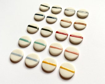 May include: A collection of 18 ceramic round buttons with a single colored stripe across the center. The buttons are arranged in a triangular pattern with the colors ranging from black, navy, dark blue, brown, red, pink, and yellow.