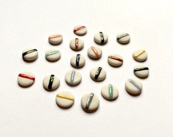 May include: A collection of 17 small, round ceramic tiles with a white background and a single colored stripe across the center. The stripes are in various colors, including red, orange, yellow, green, blue, and black.
