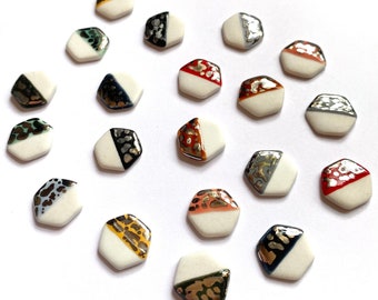May include: A collection of small, white, hexagonal ceramic beads. Each bead has a diagonal section with a metallic or patterned design in colors like gold, silver, red, and black. The beads are scattered on a white surface.