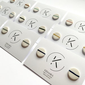 May include: A set of ceramic stud earrings with white bases and coloured stripes. The earrings are arranged on white cards with the brand name "KERAMIKA" printed on them. The brand name is also printed on the cards in a stylised font.
