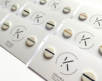 May include: A set of ceramic stud earrings with white bases and colored stripes. The earrings are arranged on white cards with the brand name "KERAMIKA" printed on them. The brand name is also printed on the cards in a stylized font.