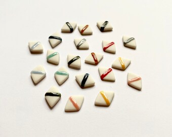 May include: A collection of small, white, diamond-shaped ceramic tiles. Each tile features a diagonal stripe in various colors, including black, silver, red, and yellow. The tiles are arranged on a white surface, creating a visually appealing pattern.
