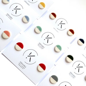 Könnte beinhalten: Sortiment an Keramik-Halbkreis-Ohrringen in verschiedenen Farben, darunter Rot, Blau, Grün, Gelb, Braun und Grau. Jeder Ohrring hat einen weißen Halbkreis an der Unterseite. Die Ohrringe werden auf weißen Karten mit dem aufgedruckten Text "KERAMIKA made in manchester" präsentiert.