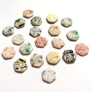 May include: A collection of 18 hexagonal ceramic tiles with various colourful paint splatters. The tiles are arranged in a circular pattern on a white background.