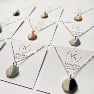 Könnte beinhalten: Ein Set aus sechs silbernen Halsketten mit sechseckigen Keramik-Anhängern. Die Anhänger sind weiß mit einem einzigen Farbakzent auf einer Hälfte. Die Farben sind grün, rosa, orange, gelb, blau und schwarz.