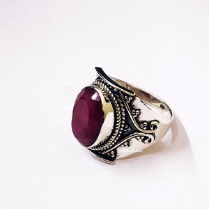 Op de afbeelding: Een zilveren ring met een ovale robijn-edelsteen gezet in een gedetailleerde zilveren setting. De ring heeft een getextureerde band en ingewikkelde ontwerpen.