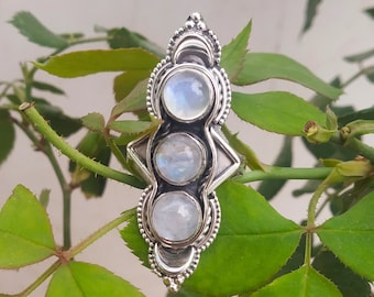 May include: A silver ring with three moonstone gems set in a decorative design. The ring is on a green leafy background.