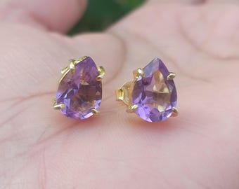 May include: A pair of gold stud earrings featuring pear-shaped, purple amethyst gemstones. The earrings are set in a gold metal with a four-prong setting, showcasing the gemstones' facets. The earrings are displayed on a light pink surface.