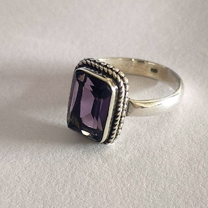 Peut inclure: Une bague en argent avec une pierre précieuse violette sertie dans un bézel carré. Le bézel est orné d'un motif de corde autour du bord.