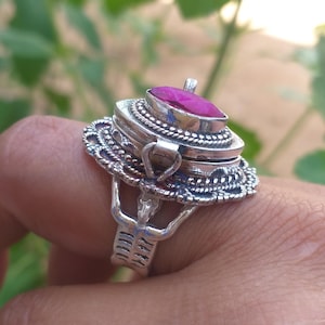 Op de afbeelding: Een zilveren ring met een scharnierend compartiment en een roze edelsteen bovenop. De ring is versierd met ingewikkelde details.