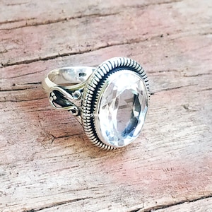 Può includere: Un anello d'argento con una grande pietra preziosa trasparente sfaccettata incastonata in una montatura a castone. L'anello ha una fascia dettagliata e decorata.
