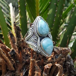 Puede incluir: Un anillo de plata con dos piedras preciosas de color azul claro en forma de lágrima. El anillo tiene un diseño intrincado y detallado con un borde trenzado y elementos decorativos. El anillo se encuentra sobre un fondo natural de hojas verdes y ramas marrones.