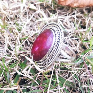 May include: A silver ring with a large, oval-shaped, red gemstone set in a silver bezel. The ring has a decorative, braided band.