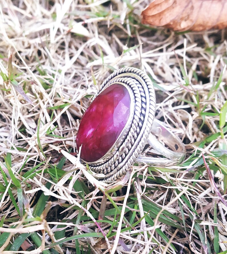 May include: A silver ring with a large, oval-shaped, red gemstone set in a silver bezel. The ring has a decorative, braided band.