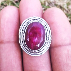 May include: A silver ring with a large, oval-shaped, deep pink ruby gemstone set in a decorative, rope-like border.
