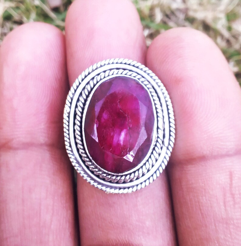 May include: A silver ring with a large, oval-shaped, deep pink ruby gemstone set in a decorative, rope-like border.