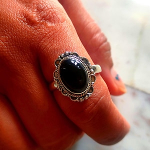 Könnte beinhalten: Ein silberner Ring mit einem ovalen schwarzen Edelstein, der in einem dekorativen Filigranmuster gefasst ist.