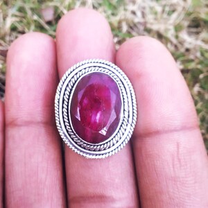 May include: A silver ring with a large, oval-shaped ruby gemstone. The ruby is a deep red color and has a faceted cut. The ring has a simple, elegant design.