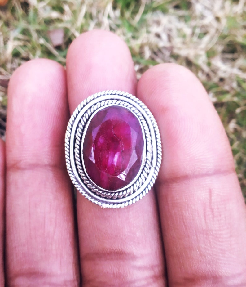 May include: A silver ring with a large, oval-shaped ruby gemstone. The ruby is a deep red color and has a faceted cut. The ring has a simple, elegant design.