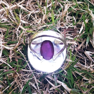 May include: A silver ring with a large, deep purple gemstone set in the center. The ring is surrounded by a decorative silver band.