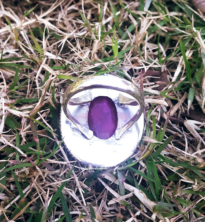 May include: A silver ring with a large, deep purple gemstone set in the center. The ring is surrounded by a decorative silver band.