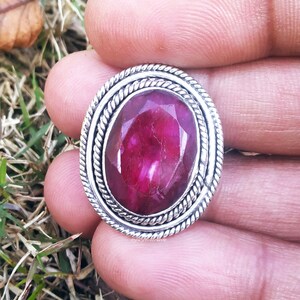 May include: A silver ring with a large, oval-shaped, dark pink gemstone set in a silver bezel. The ring has a decorative, twisted rope design around the bezel.