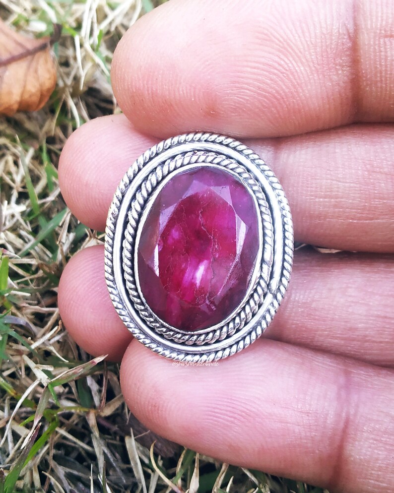 May include: A silver ring with a large, oval-shaped, dark pink gemstone set in a silver bezel. The ring has a decorative, twisted rope design around the bezel.