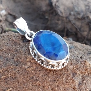 Op de afbeelding: Zilveren hanger met een ovale, diepblauwe edelsteen. De edelsteen is gezet in een decoratief zilveren montuur met een sierlijk ontwerp. De hanger heeft een kleine lus aan de bovenkant voor een ketting.