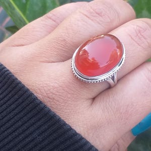 Puede incluir: Un anillo de plata con una gran piedra de cornalina de forma ovalada. La piedra es de color rojo intenso y tiene un acabado liso y pulido.