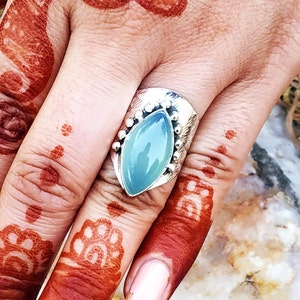 Pode incluir: Um anel de prata com uma gema azul claro engastada em uma banda texturizada. O anel é adornado com pequenas contas de prata ao redor da gema.