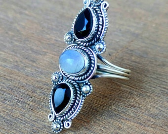 May include: A silver ring with three stones: a white moonstone and two black onyx stones. The ring has an intricate design with a beaded border.