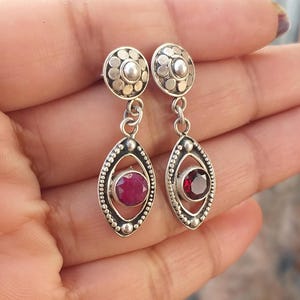 Puede incluir: Un par de pendientes de plata con una piedra preciosa roja en el centro de cada pendiente. Los pendientes tienen un diseño decorativo con un círculo en la parte superior y una forma de lágrima en la parte inferior.