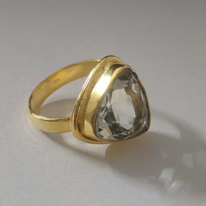 Könnte beinhalten: Ein goldener Ring mit einem großen, klaren, dreieckigen Edelstein. Der Ring ist in einer goldenen Fassung mit einem erhöhten Rand gefasst.