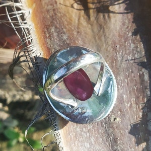 May include: A silver ring with a large, oval-shaped, dark pink gemstone set in the center. The ring is on a brown, textured surface.