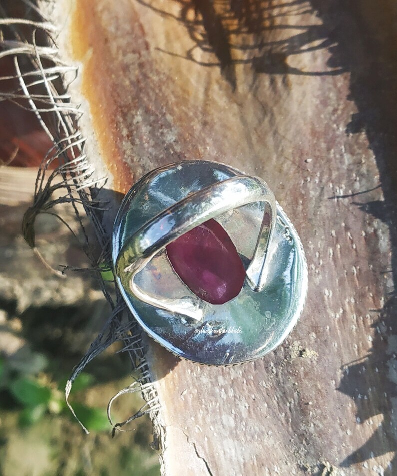 May include: A silver ring with a large, oval-shaped, dark pink gemstone set in the center. The ring is on a brown, textured surface.