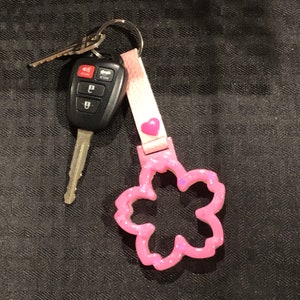 Könnte beinhalten: Ein Satz Autoschlüssel an einem rosa blütenförmigen Schlüsselanhänger befestigt. Der Schlüsselanhänger hat einen rosa Riemen mit einem kleinen rosa Herz. Der blütenförmige Anhänger ist glitzernd und hat lila Akzente.