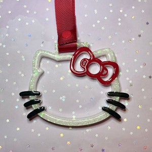 Puede incluir: Un llavero blanco brillante con forma de Hello Kitty, con un lazo rojo y bigotes negros. El llavero tiene una correa roja con cierre de botón a presión. El diseño está delineado en blanco y relleno de purpurina iridiscente.