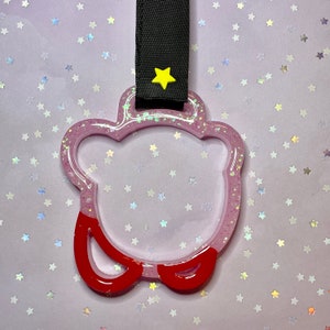 May include: A pink, glitter-filled, bear-shaped keyring with red accents. The keyring has a clear centre and a black strap with a yellow star. The background is a light purple with star-shaped confetti.