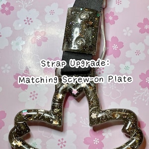 May include: A black strap upgrade with a matching screw-on plate. The plate and handle feature a clear resin with glitter and floral accents. The text "Strap Upgrade: Matching Screw-on Plate" is visible.