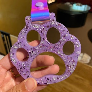 May include: A lavender paw-shaped keychain with silver glitter accents. The keychain has a holographic strap and button closure. The paw print design features four circular cutouts and is held by a hand.