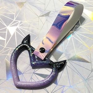 May include: A heart-shaped keychain with a gradient of purple and black glitter. The keychain has a holographic strap attached with a black snap. The design features pointed tips on the heart shape.