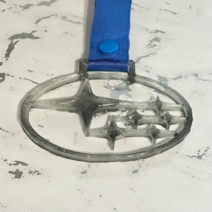 May include: A clear, oval pendant with a Subaru logo, attached to a blue ribbon with a button. The pendant has a glittery, grey interior, and the logo is a stylized star cluster. The blue ribbon is attached to the pendant with a blue button.