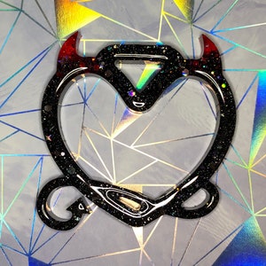 May include: Black heart-shaped charm with devil horns and a tail. The horns are red and fade into black. The charm is filled with glitter and small star-shaped accents. The background is a geometric pattern with iridescent colours.
