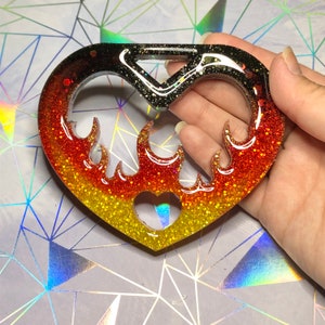 May include: A heart-shaped keychain with a flame design. The keychain is made of glittery resin, with colors that transition from yellow to orange to black. A small heart cutout is at the bottom.