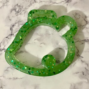 May include: A green leaf-shaped keychain made of resin, filled with glitter. The keychain has a loop at the top and a rounded shape at the bottom. The glitter is in various shades of green and gold.
