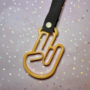 May include: A tan-colored plastic hand-shaped accessory with a black strap and a white button. The hand is in a gesture with the index and middle fingers extended. The background is a light purple color with star-shaped glitter.