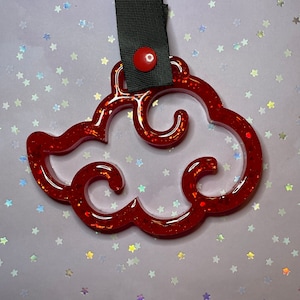 Può includere: Ciondolo a forma di nuvola rossa con glitter, attaccato a un cinturino nero con un bottone rosso. Il ciondolo è un design a nuvola stilizzato, probabilmente per un portachiavi o un accessorio. Lo sfondo è chiaro con glitter a forma di stella.