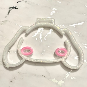 May include: A white, glitter-infused hair clip in the shape of a cartoon character's face, featuring pink accents for the cheeks. The clip has a rectangular clasp and curved edges, creating a playful aesthetic.