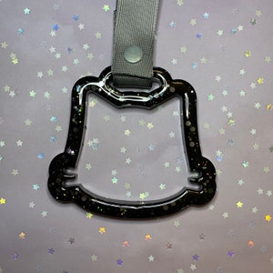 May include: A black, glitter-infused resin charm with a cat-like silhouette, featuring a gray strap and a snap closure. The charm is designed with a hollow center and is set against a background of scattered star-shaped confetti.
