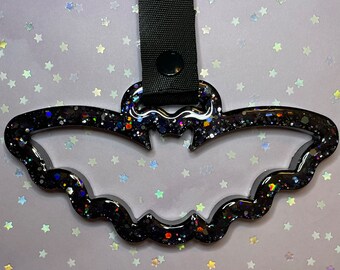 May include: Black bat-shaped keychain with glitter and a black strap. The bat has a glossy finish and is filled with colorful glitter. The strap has a black snap closure.